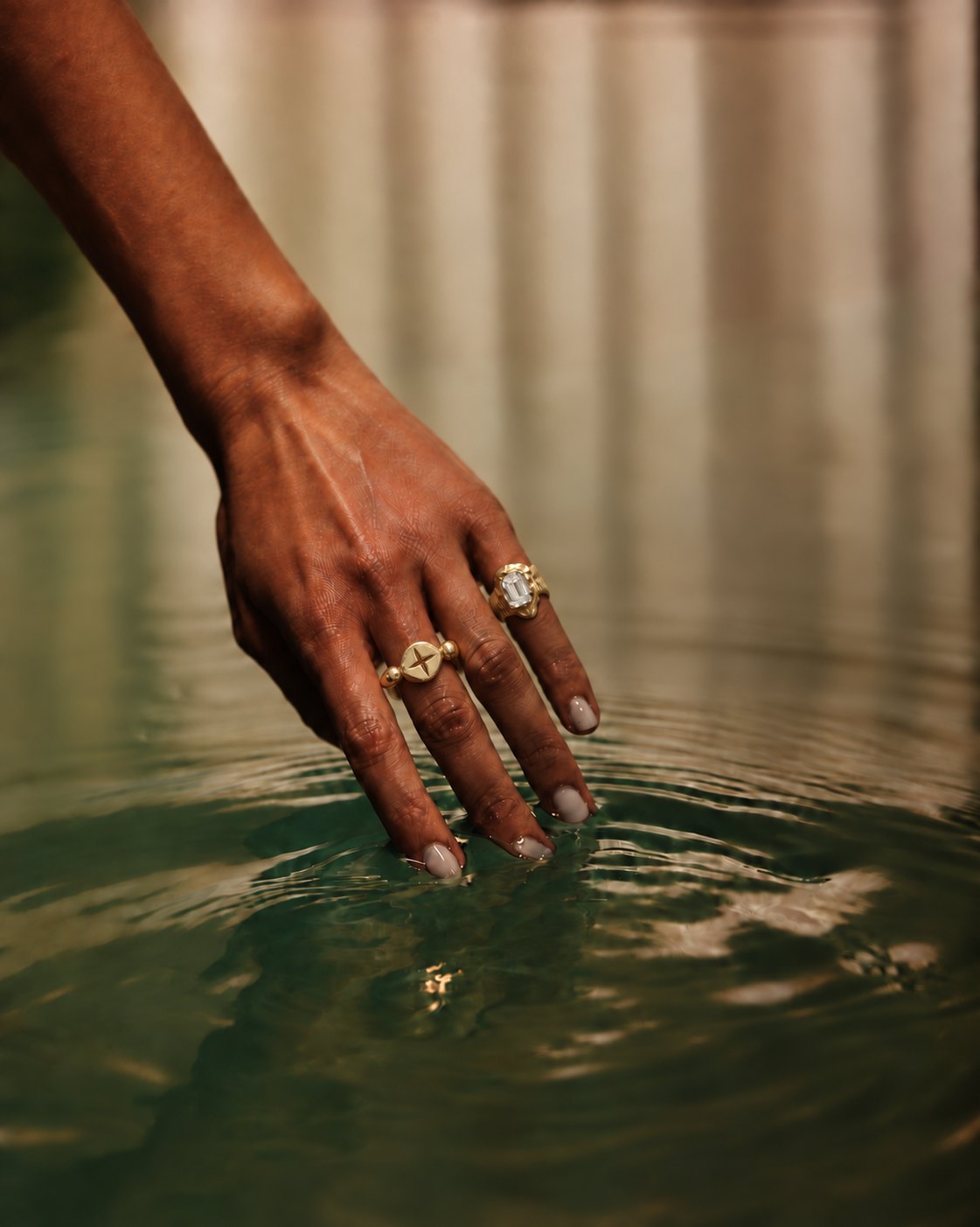 A hand wearing two SAESSO gold rings, fingertips touching still green water